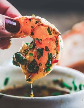 One grilled shrimp held in hand with the roasted garlic cilantro sauce dripping in the bowl