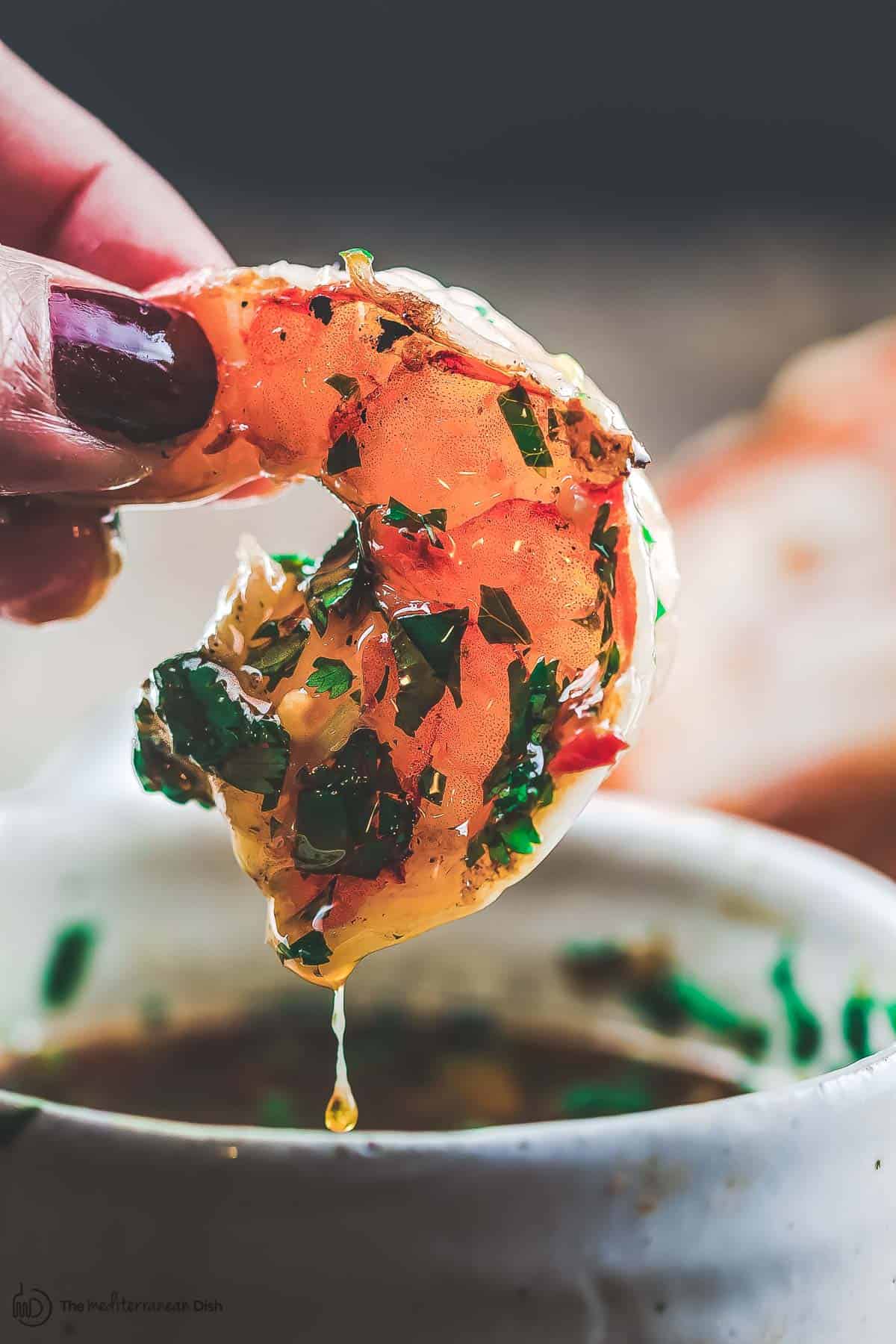 One grilled shrimp held in hand with the roasted garlic cilantro sauce dripping in the bowl