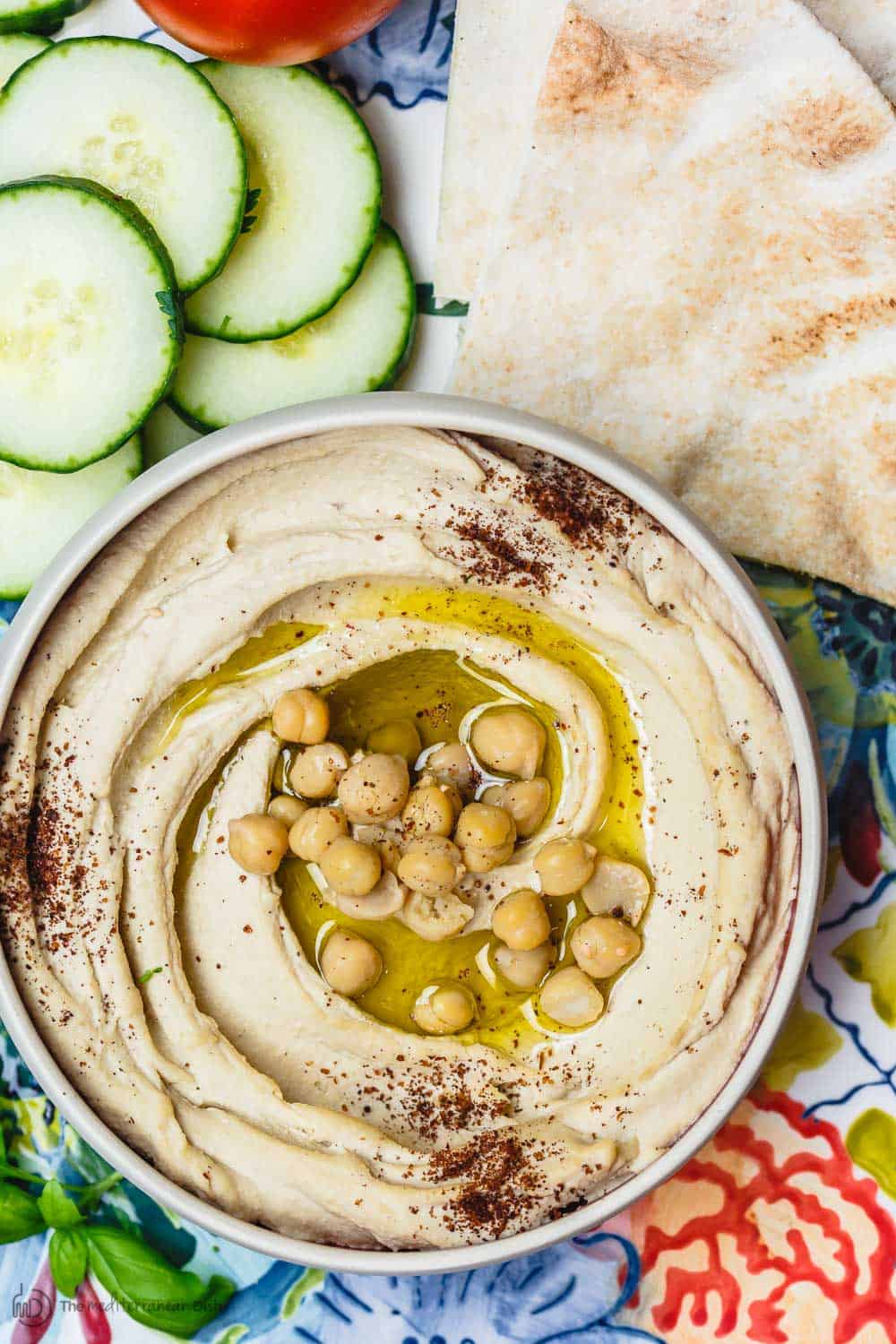 hummus served in a bowl and garnished with olive oil, chickpeas and sumac. A side of pita and cucumbers