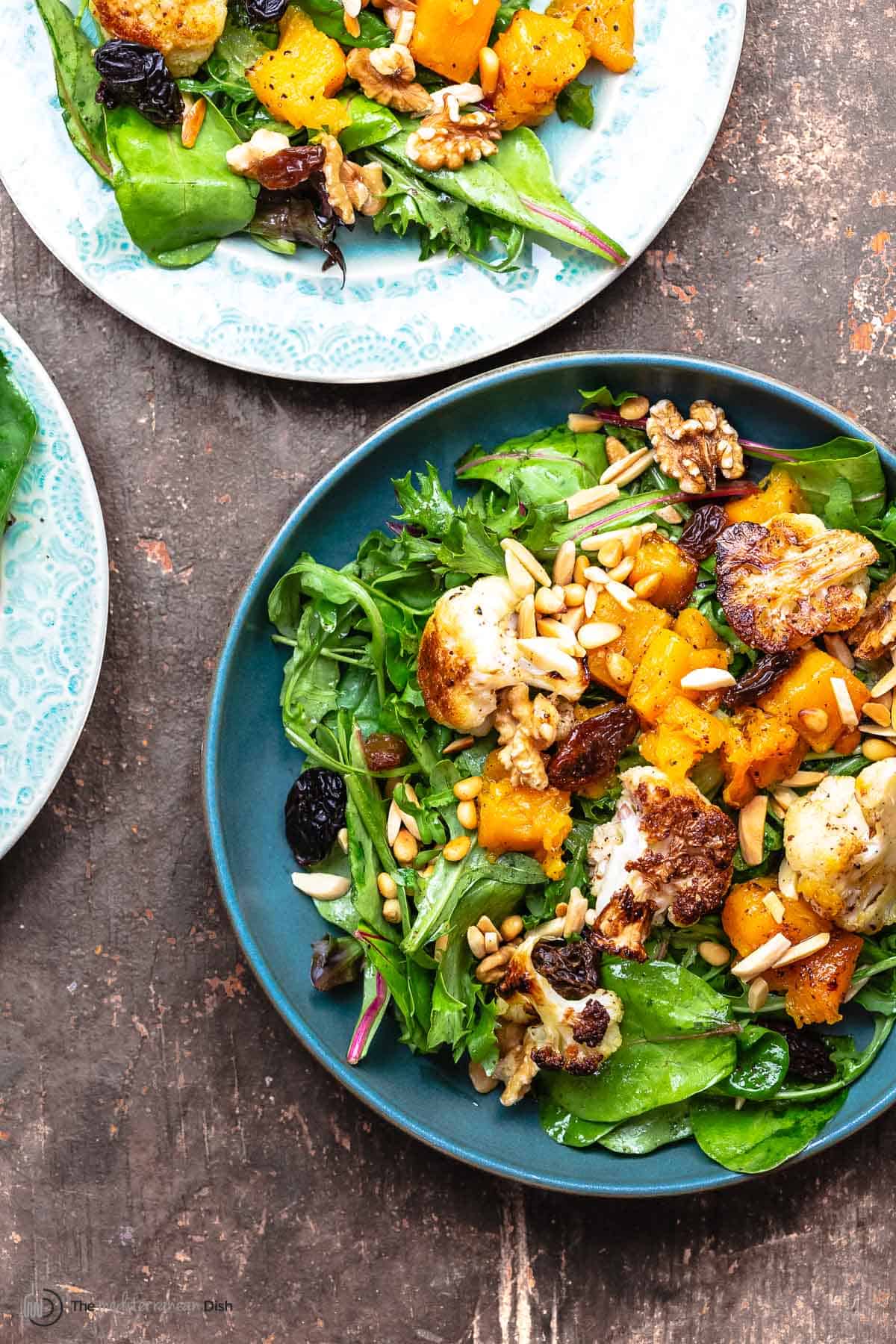 two plates with a bed of greens, roasted cauliflower and butternut salad, topped with raisins and nuts