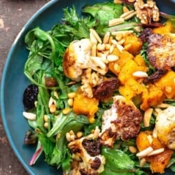 roasted cauliflower and butternut salad on a bed of greens, topped with nuts and raisins, on a blue plate