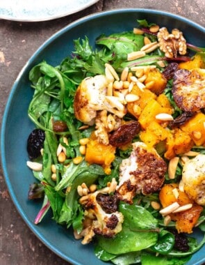 roasted cauliflower and butternut salad on a bed of greens, topped with nuts and raisins, on a blue plate