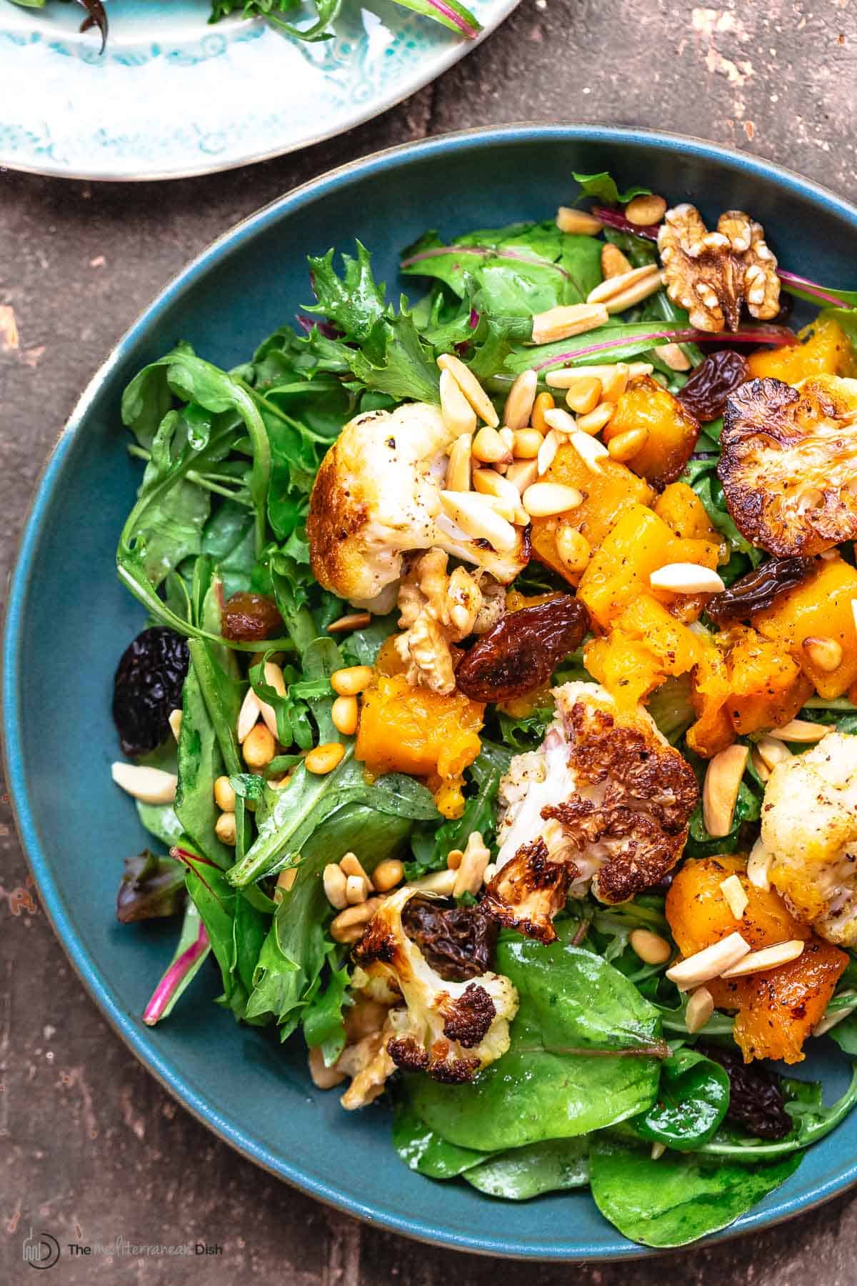 roasted cauliflower and butternut salad on a bed of greens, topped with nuts and raisins, on a blue plate