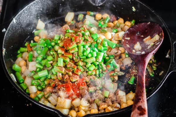 Ingredients of hash cooking in skillet. Onions, garlic, potatoes, chickpeas, asparagus, a dash more salt and pepper and the spices.