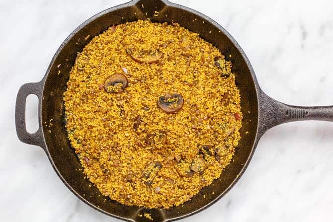 Skillet containing couscous, mushrooms, garlic, onions, spices, and the mixture of pomegranate molasses, lemon juice and olive oil.