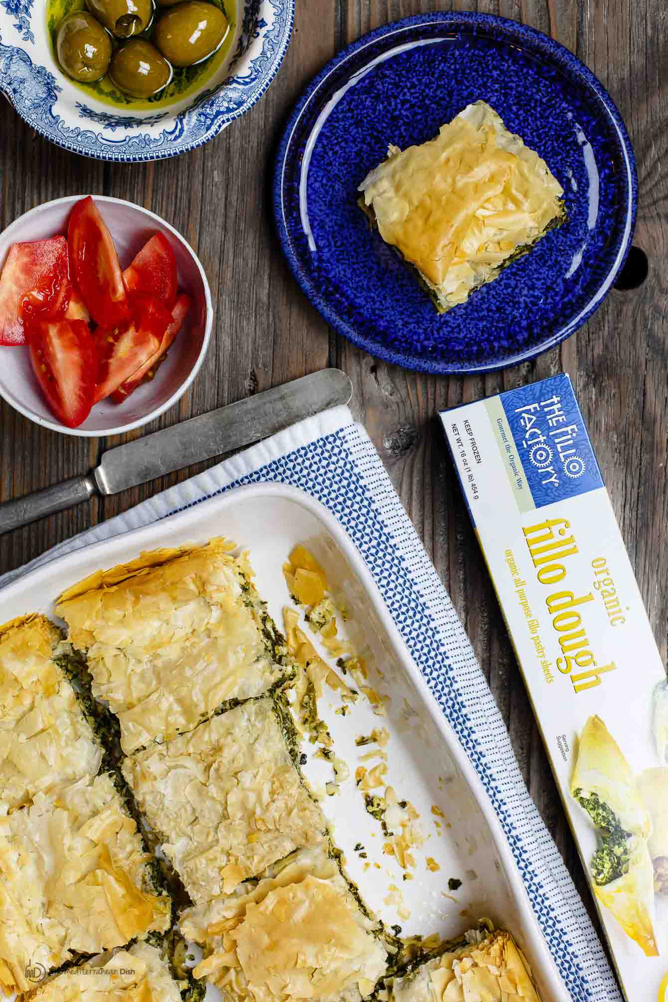 Spanakopita pie served next to olives and fresh tomatoes. Phyllo dough box on side.