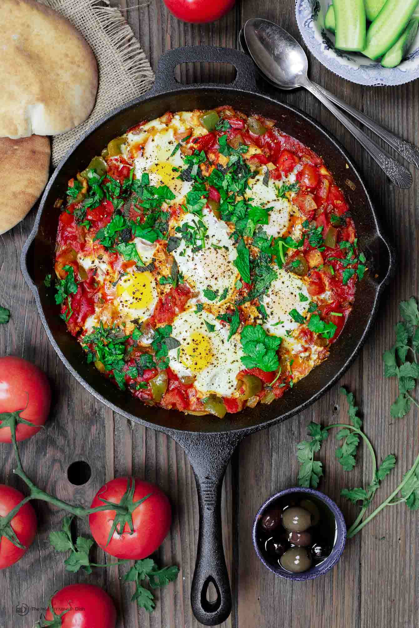 poached eggs in shakshuka tomato sauce in cast iron skillet with pita bread and sides of cucumbers and olives