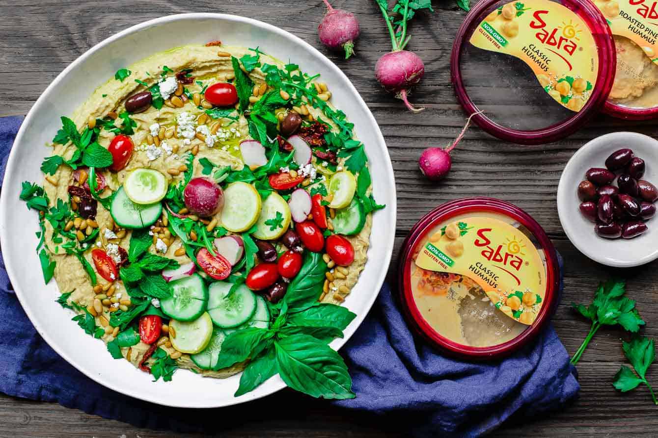 Container of Sabra dip next to bowl of hummus and veggies