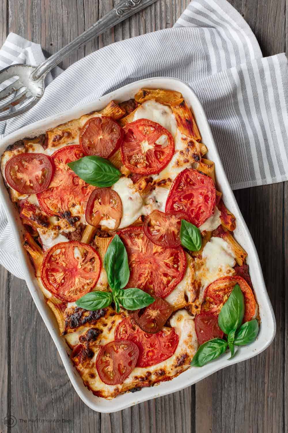 rigatoni pasta in a baking dish, topped with cheese, tomato slices and fresh basil
