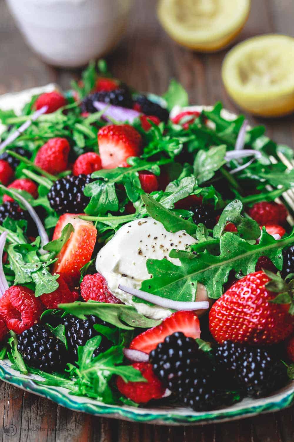 Arugula, berries, burrata and onions on a platter with a side of berry salad dressing and two lemon halves 