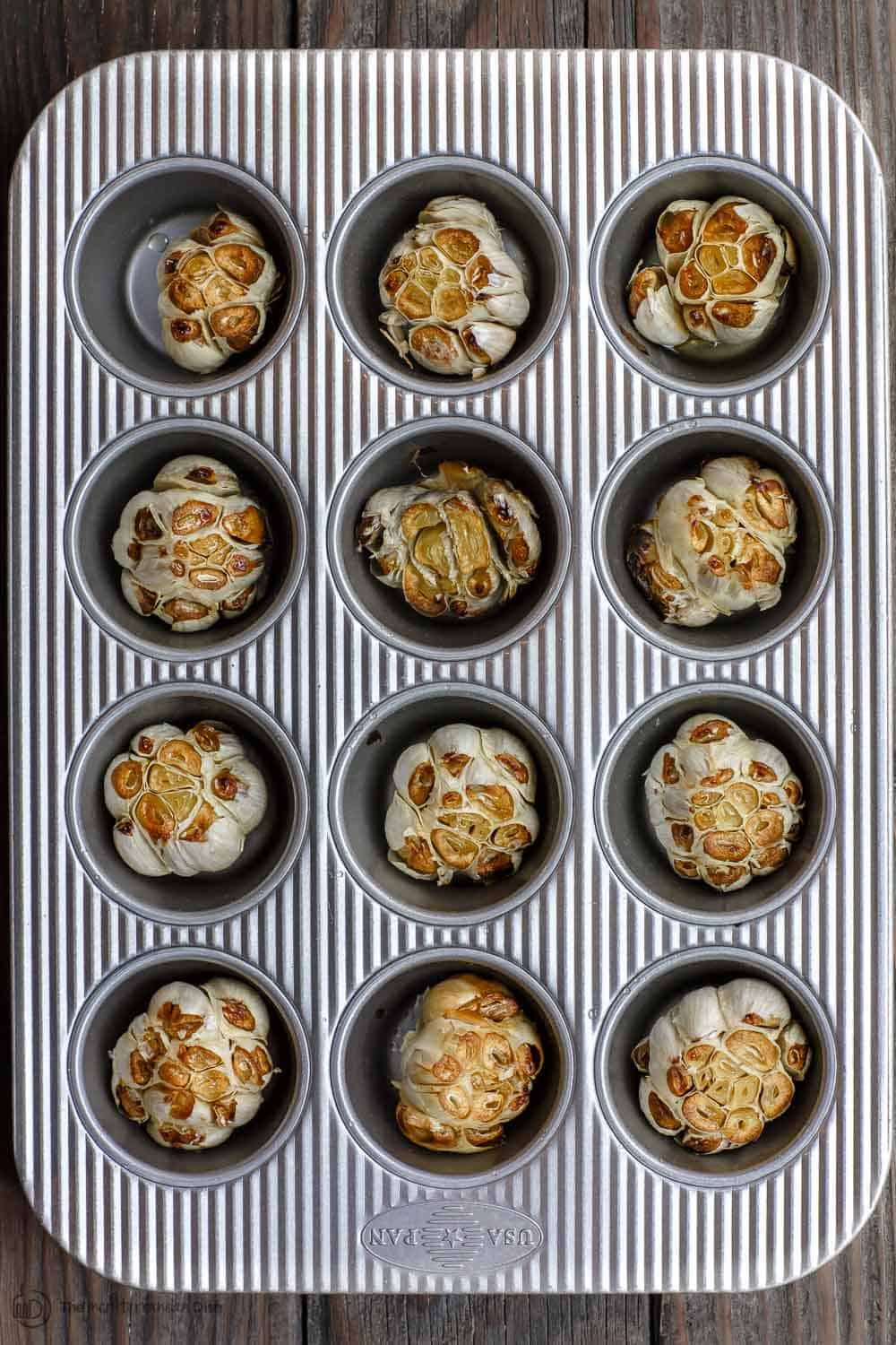 Twelve garlic heads in a muffin pan after being roasted. Perfect way to neatly and successfully roast garlic.