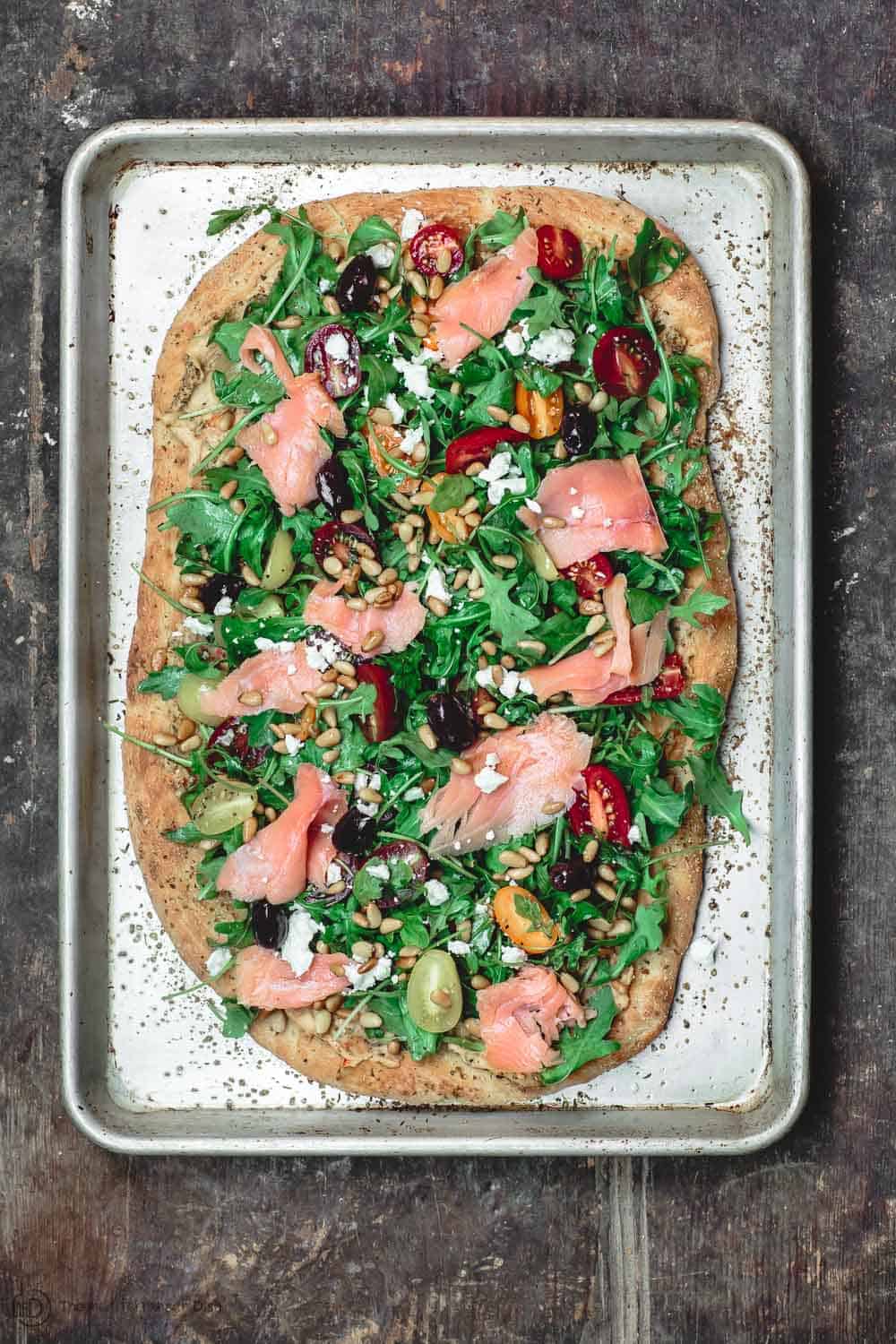 Mediterranean Flatbread served in a tray and topped with Arugula, Tomatoes, Smoked Salmon and More