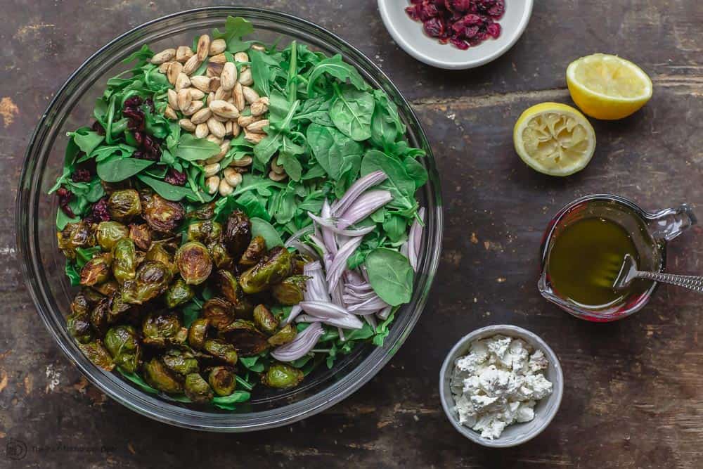 Large mixing bowl containing brussel sprouts, spinach, arugula, shallots, and almonds. A small bowl of feta cheese and a small bowl of cranberries on the side. Vinaigrette ready to be pourd