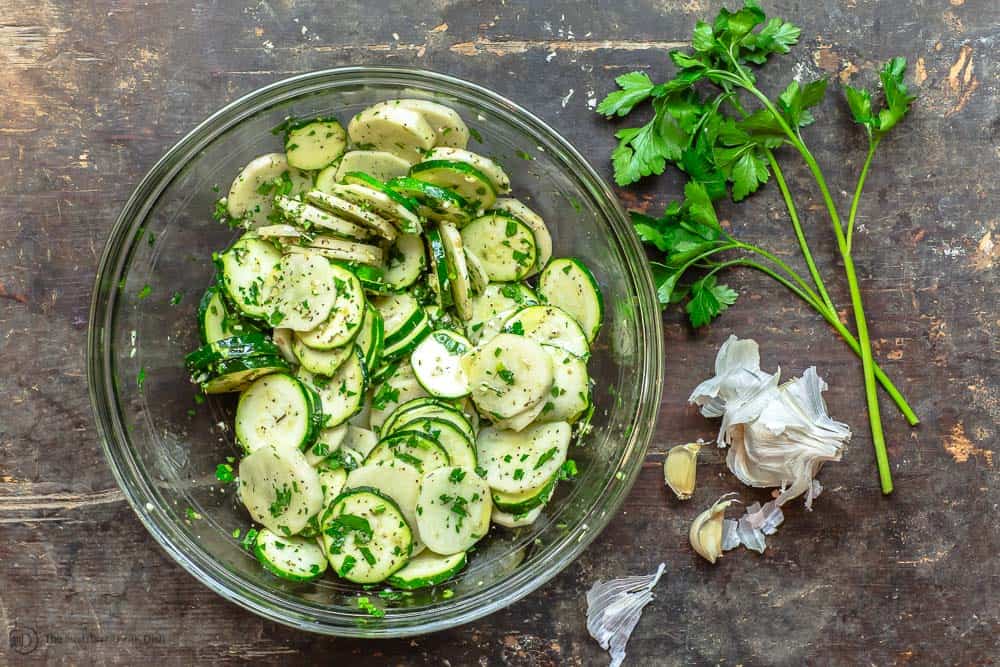 Large mixing bowl with thin slices of zucchini and potatoes tossed with spices, garlic, fresh parsley and extra virgin olive oil