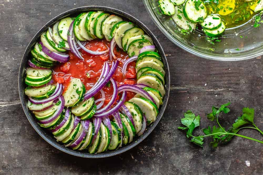 Round baking pan with diced tomato on bottom. One row of potatoes, zucchini, and red onions assembled on top. 