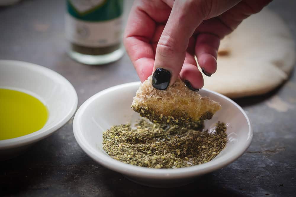 A small bowl of early harvest extra virgin olive oil and another bowl of za'atar spice. A hand holding a piece of pita bread that's been dipped in the oil then the za'atar spice