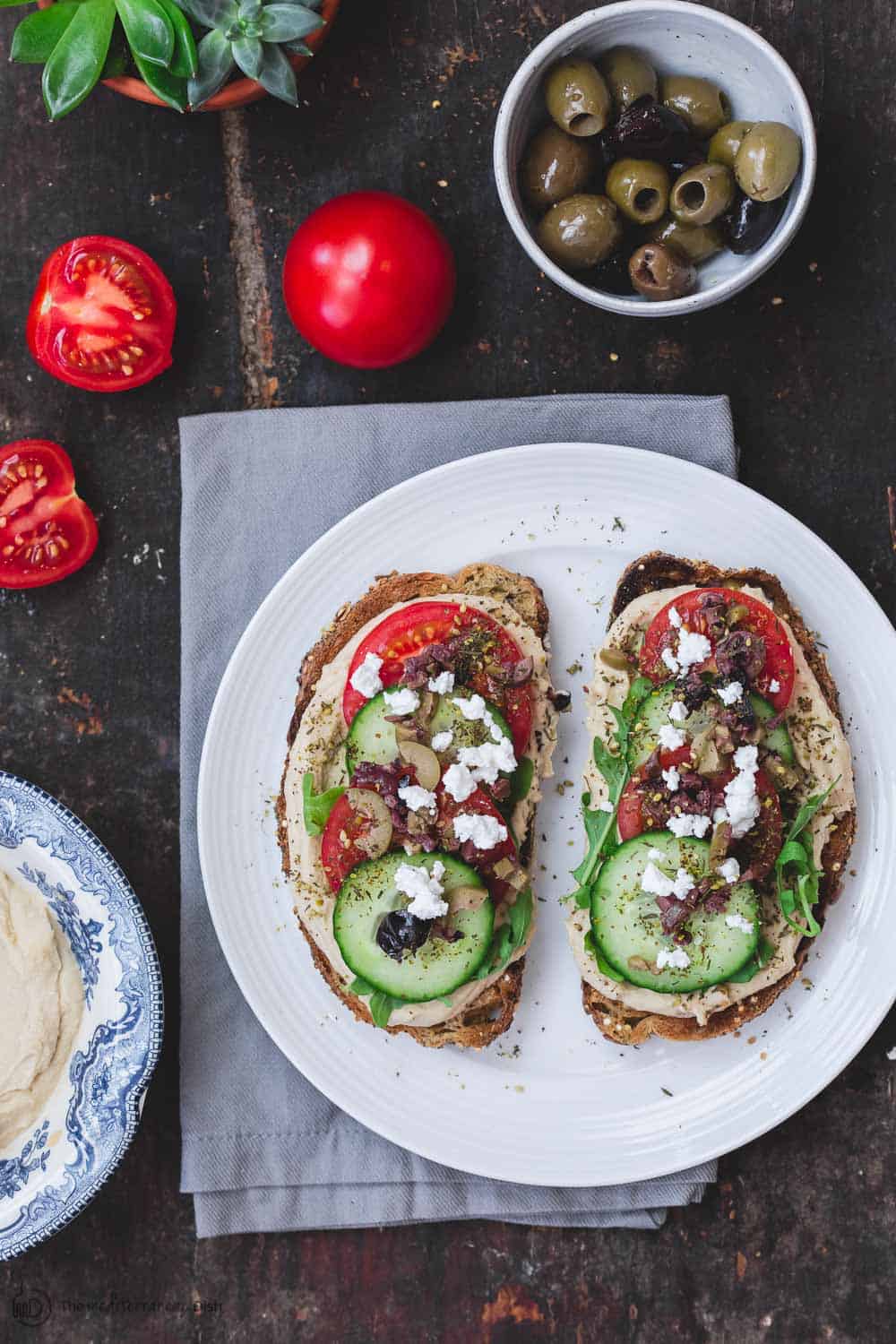 Two breakfast toasts, a bowl of olives and extra tomatoes and hummus on the side