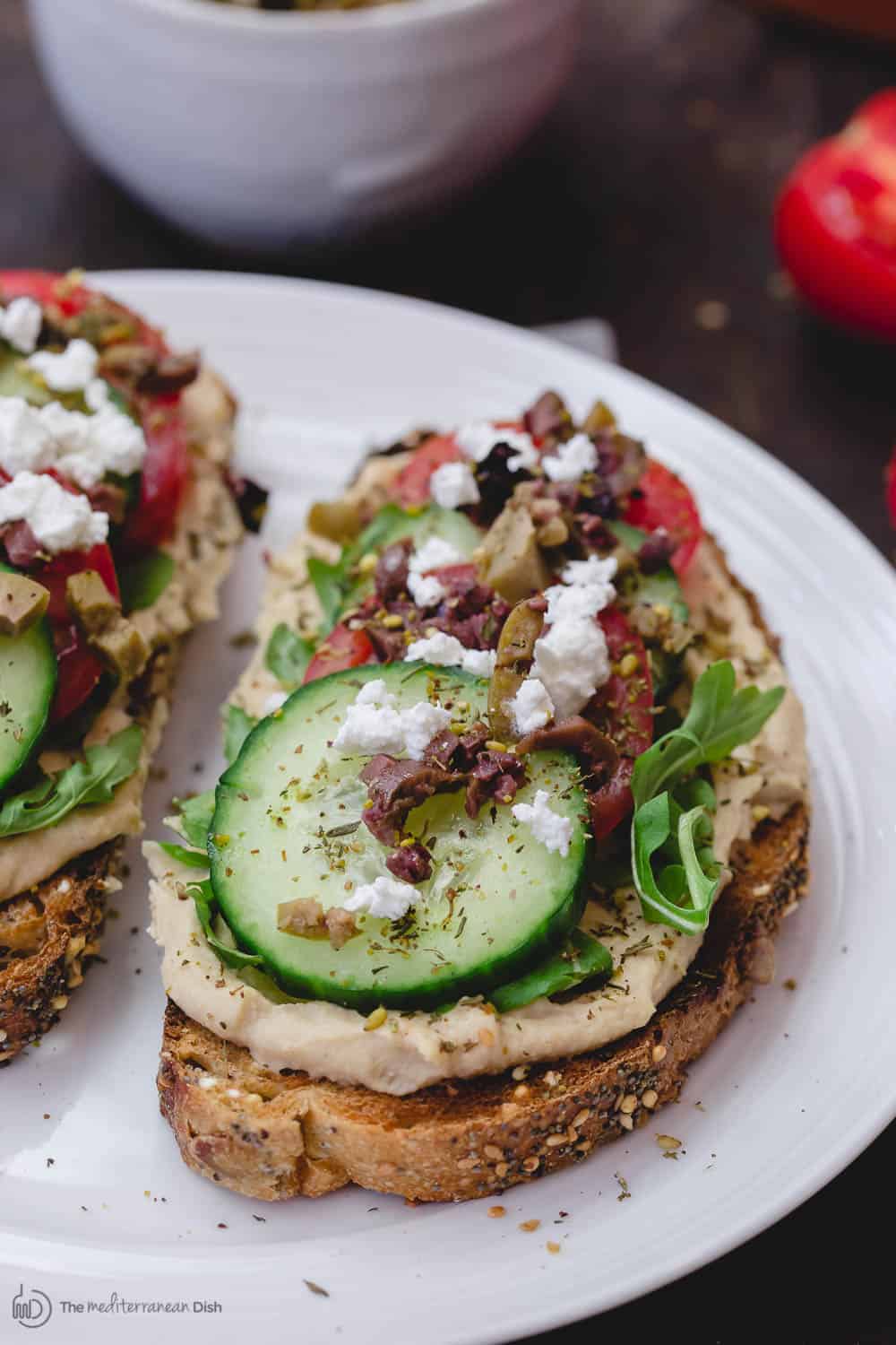 Mediterranean-Style Breakfast Toast with hummus, arugula, cucumbers, tomatoes, olives, a sprinkle of za'atar and feta cheese