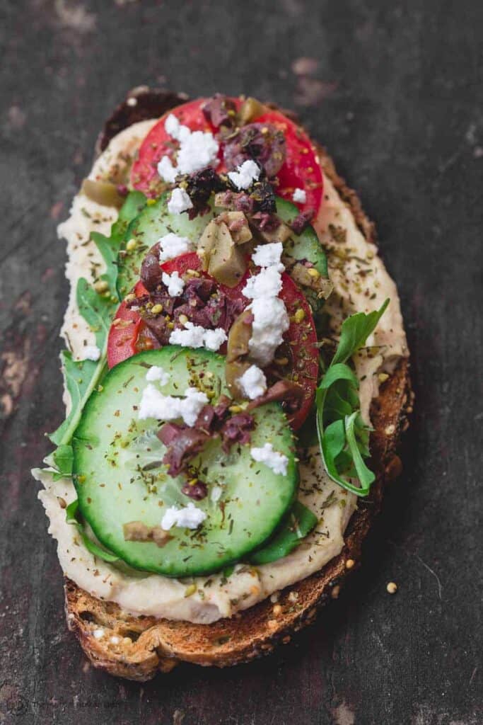 One slice of Mediterranean-style breakfast toast loaded with hummus, arugula, cucumbers, tomatoes, olives, a sprinkle of za'atar and feta cheese