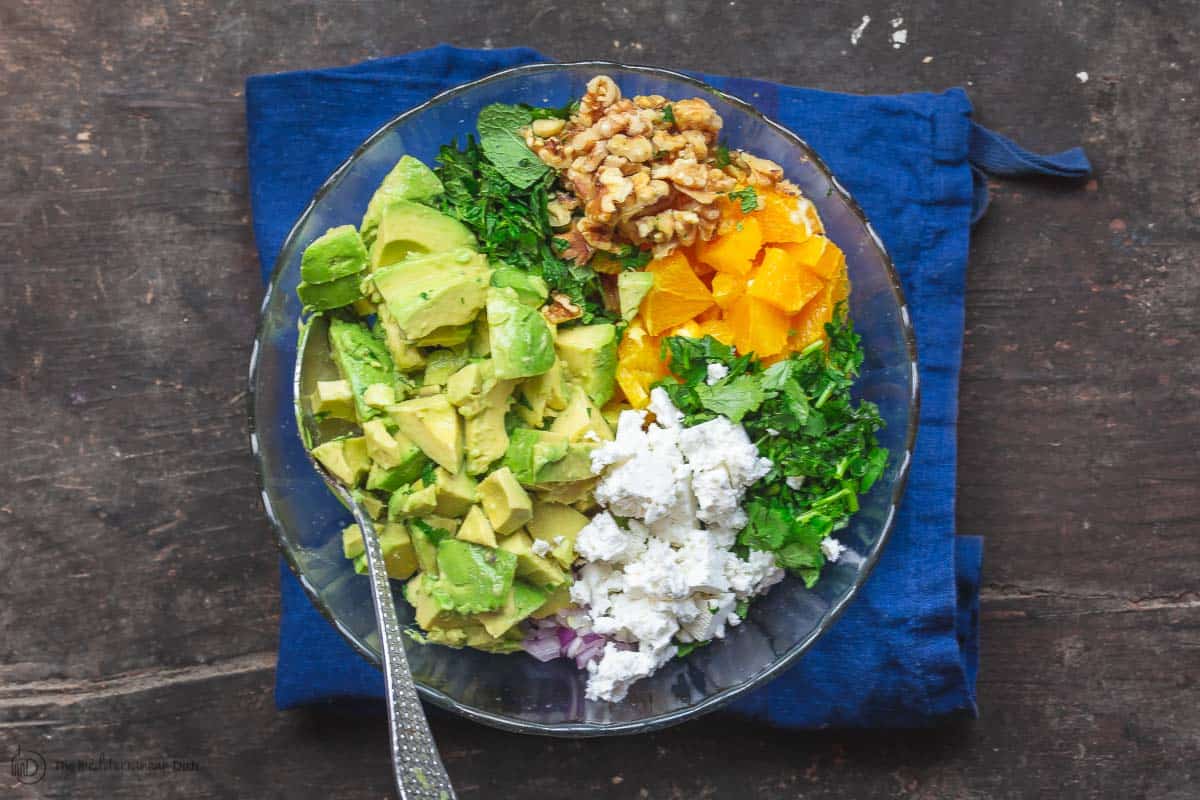 Diced avocados, feta, fresh parsley and mint, diced oranges and walnut hearts in a mixing bowl 