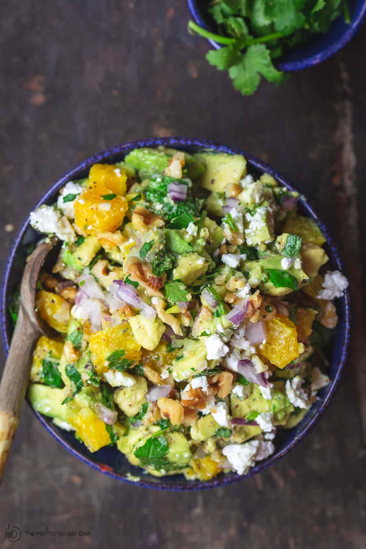 Avocado salsa served in a blue bowl with orange, walnuts, feta, and fresh herbs