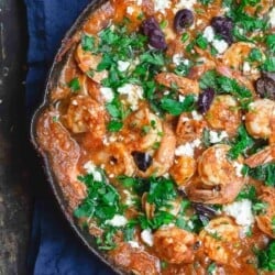 Greek shrimp in skillet, doused in tomato sauce and topped with chopped fresh herbs, crumbled feta cheese, and olives