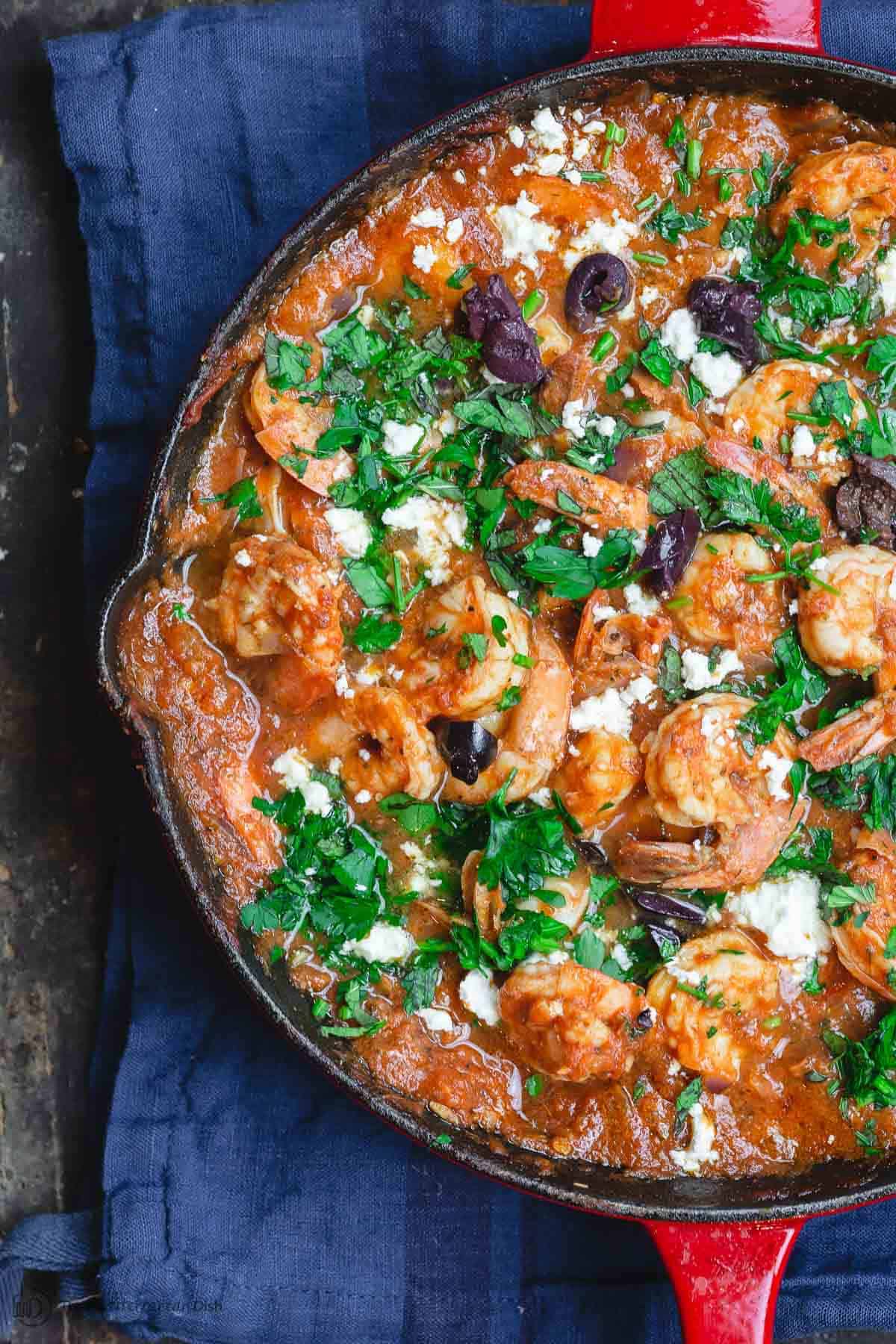 Greek shrimp in skillet, doused in tomato sauce and topped with chopped fresh herbs, crumbled feta cheese, and olives
