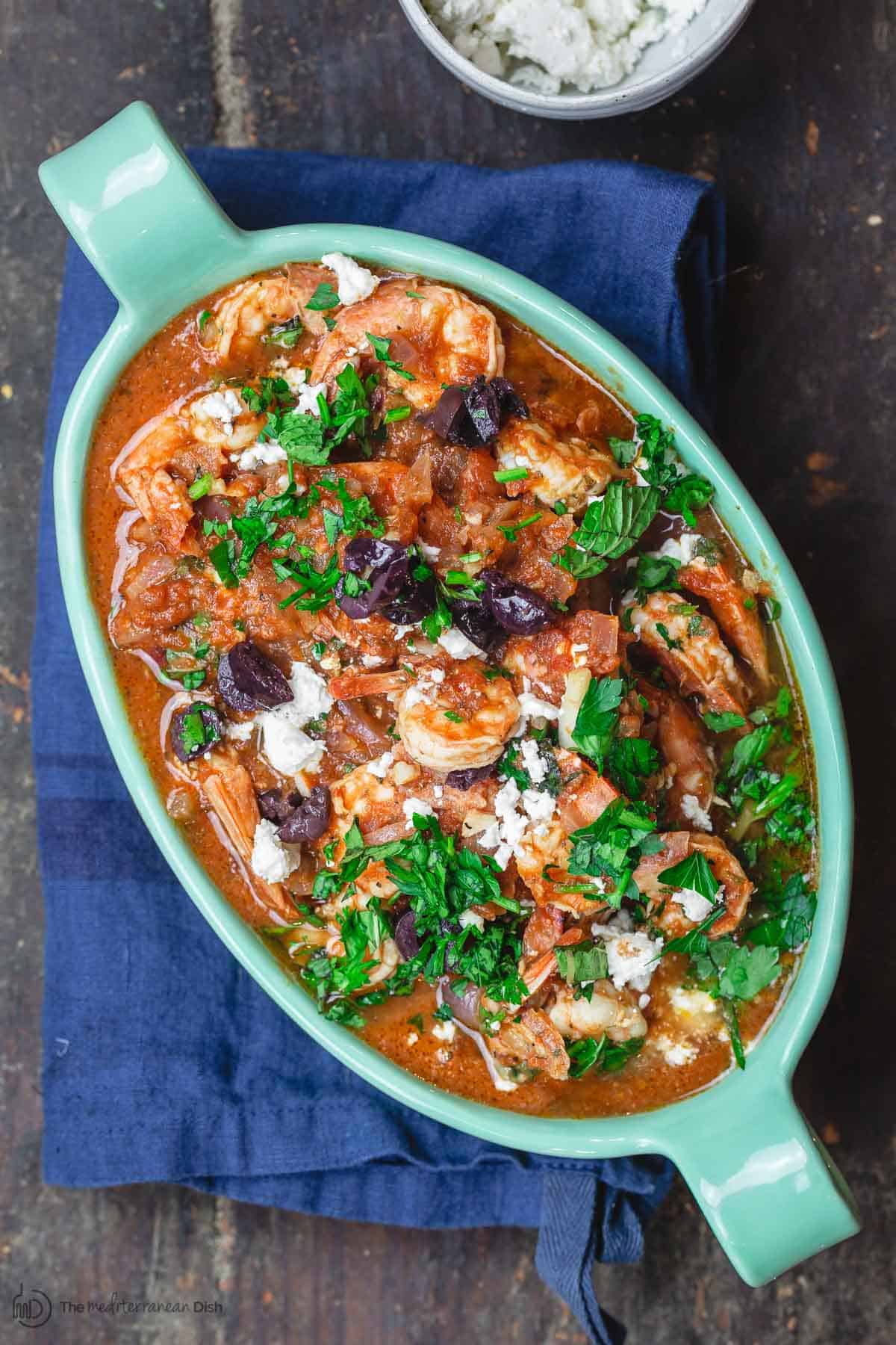 Greek shrimp in tomato sauce, served in deep serving dish and topped with fresh chopped herbs, feta cheese, and kalamata olives. A small bowl of feta cheese to the side.