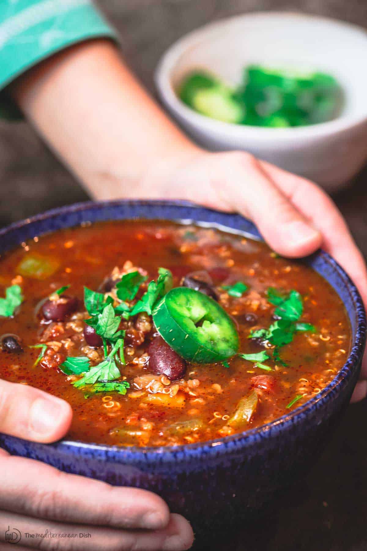 Dinner bowl of vegan chili held in two hands as it's placed on table. Topping the bowl of chili are some beans, fresh herbs and sliced jalapeno.