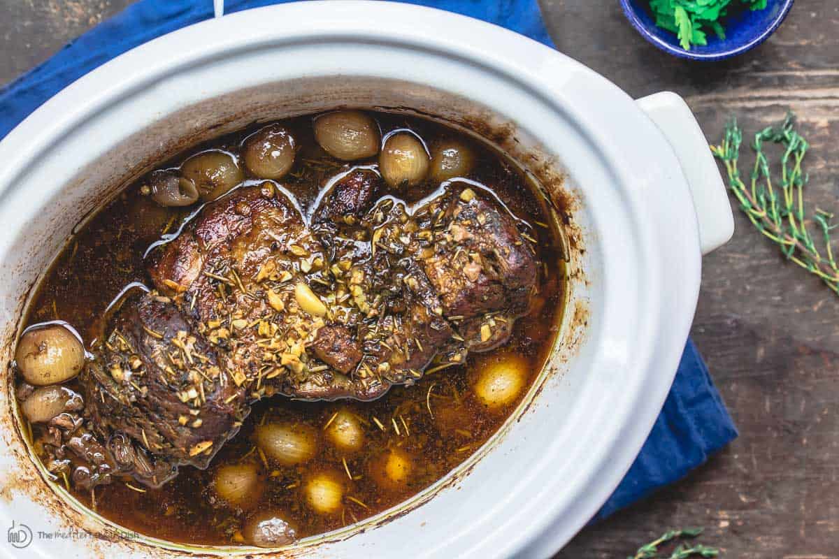 Boneless leg of lamb in slow cooker with pearl onion and wine broth. Fresh herbs to the side for garnish