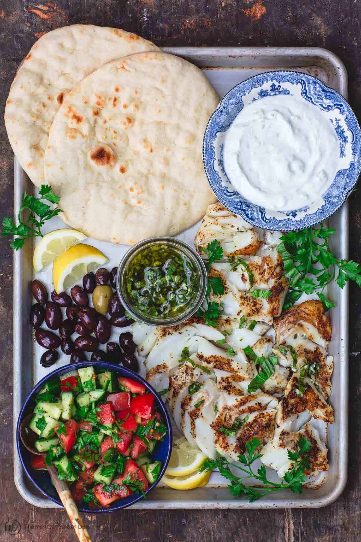 Mediterranean Pan Grilled Cod served on a tray with lemon basil sauce and gyro fixings like pita, salad, olives and Tzatziki