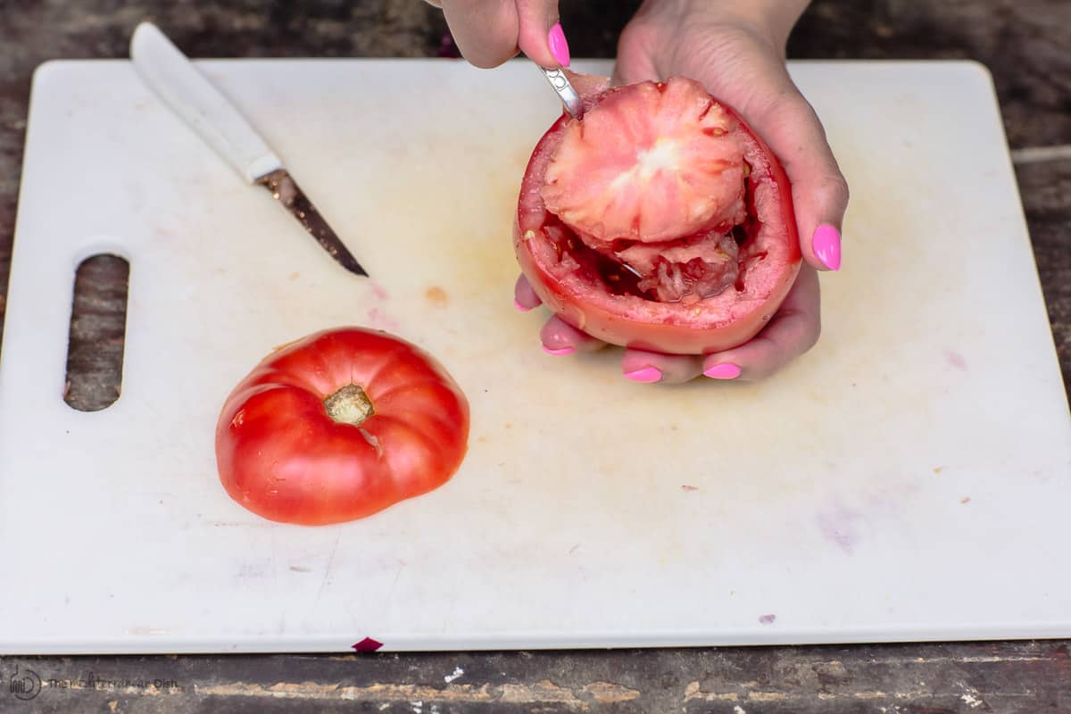 A spoon is used to carefully scoop up tomato flesh, preparing for stuffed tomatoes