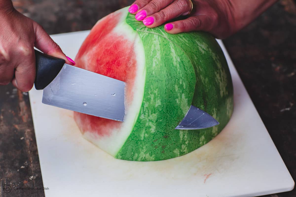 A watermelon half on a cutting board and a person's hand holding a knife and removing the rind