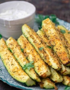 Baked zucchini sticks, stacked on a plate with a side of Tzatziki sauce