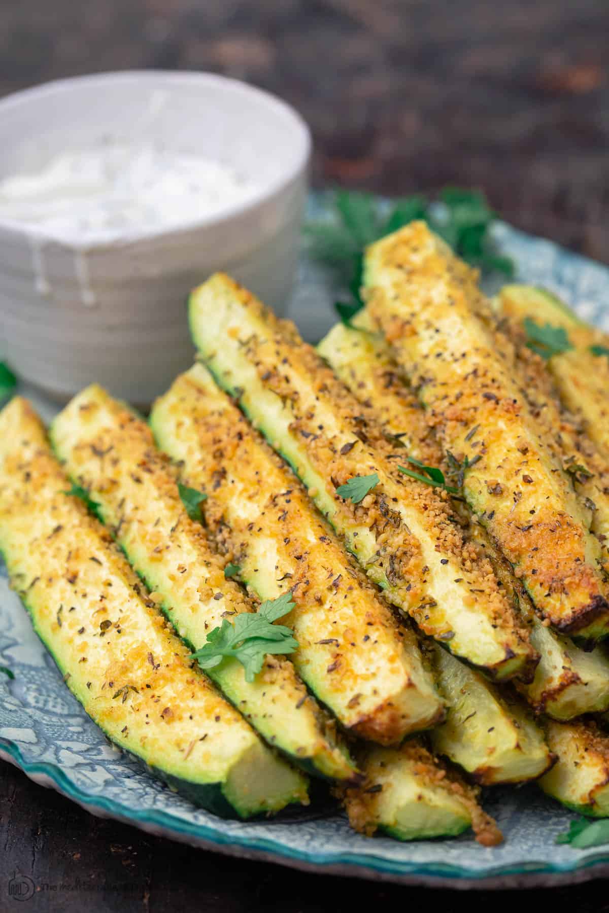 Baked zucchini sticks, stacked on a plate with a side of Tzatziki sauce