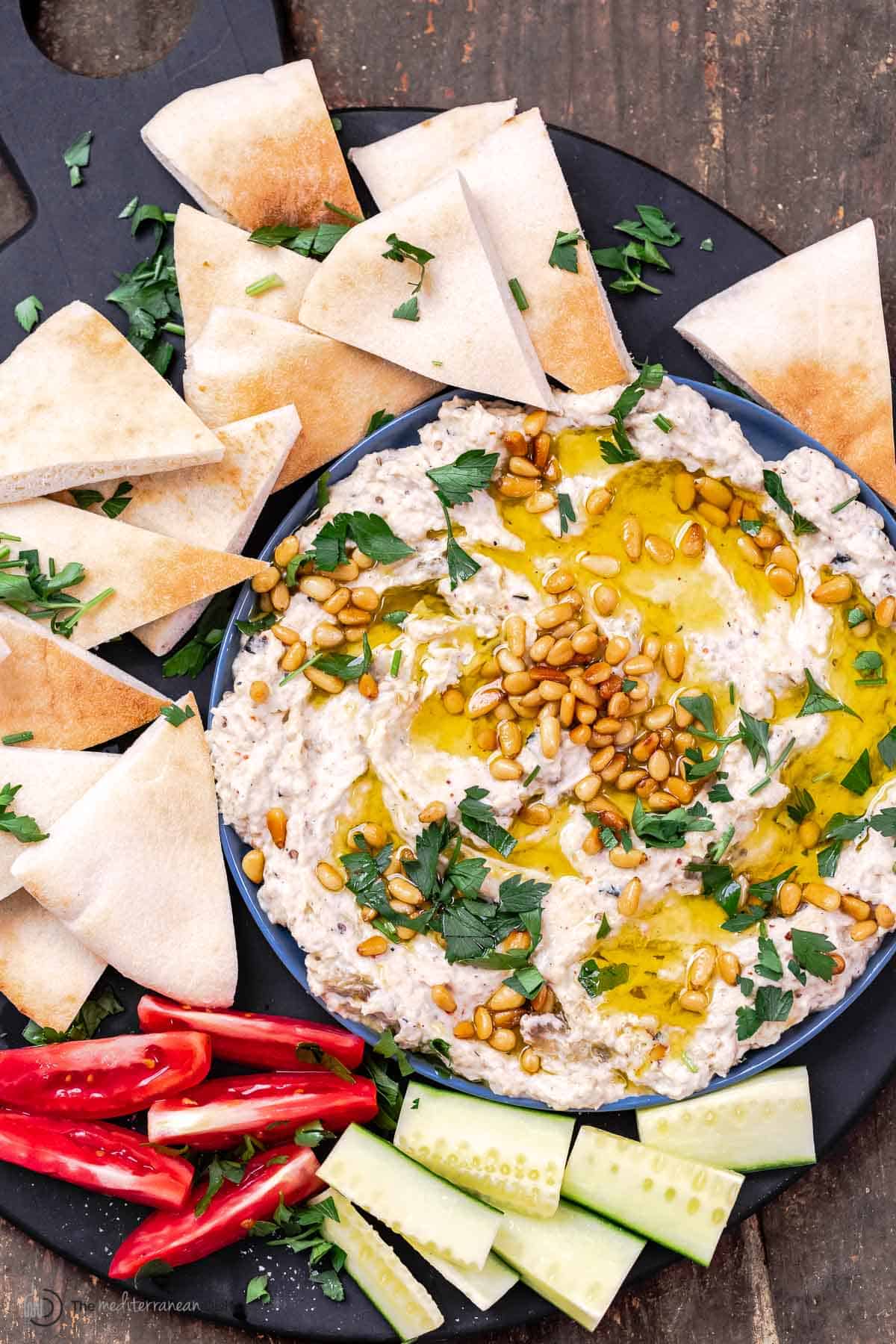 Baba ganoush dip topped with olive oil and toasted nuts and served with bell peppers, cucumbers and pita bread wedges