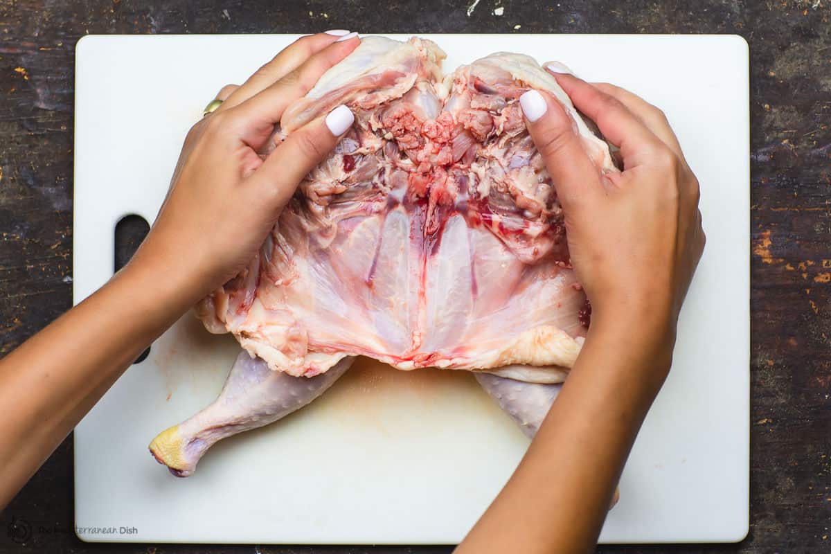 Hands pressing down on both sides of whole chicken to spread the chicken out