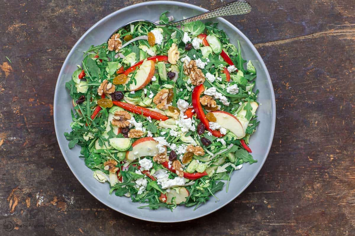 Shaved brussel sprouts salad served in large bowl, topped with feta, walnuts, cranberries and raisins