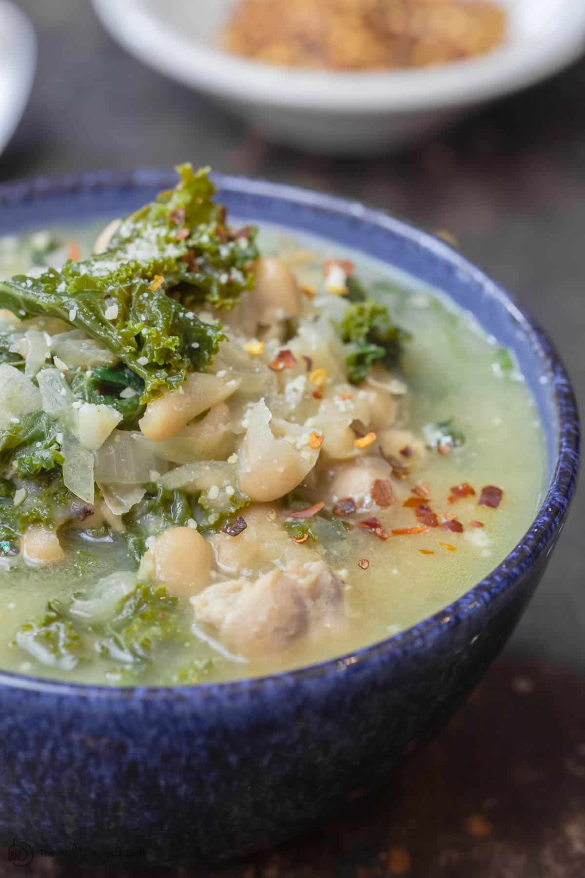 White Bean and Kale Soup served in bowl. A small bowl of red pepper flakes to the side