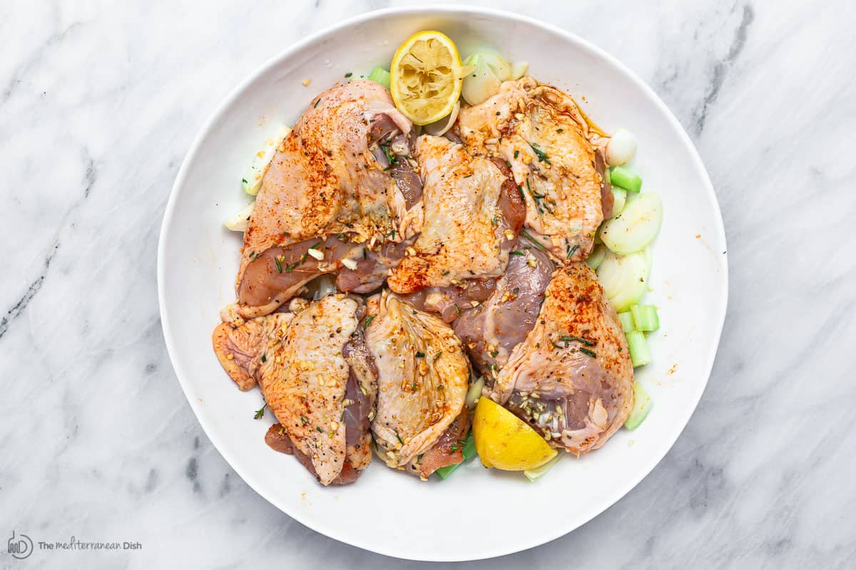 Seasoned chicken being marinated in a large bowl with extra virgin olive oil and citrus with onions and celery