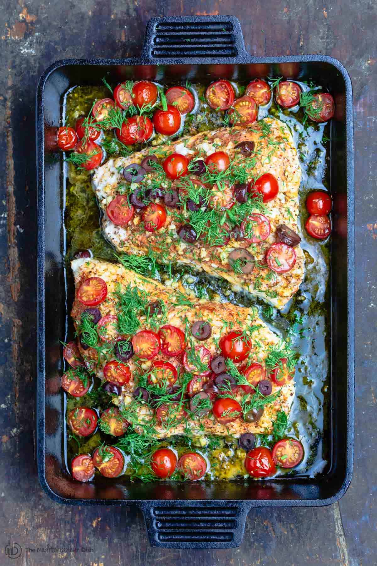 Baked grouper in black baking pan, topped with tomatoes, olives and fresh dill