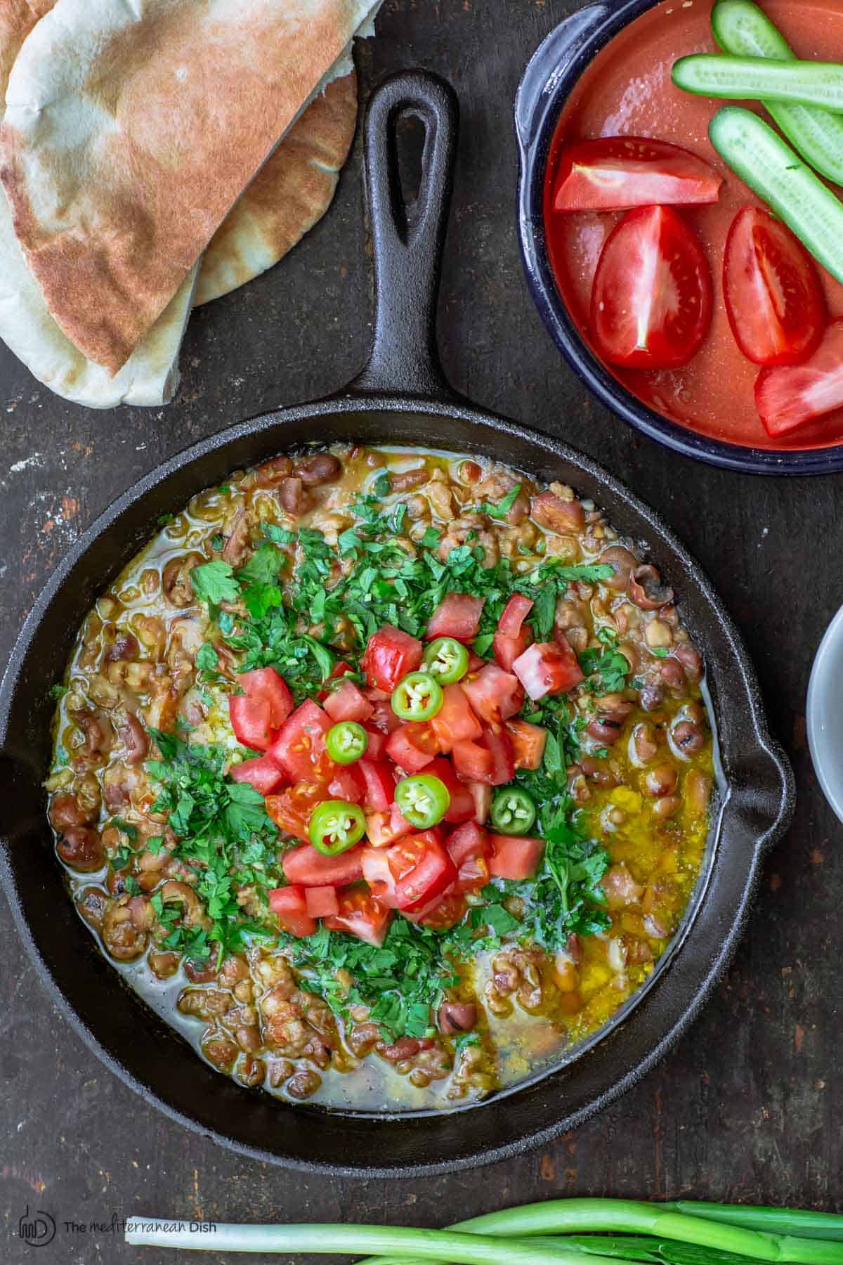 Foul Mudammas served with sides of warm pita bread, sliced vegetables and green onions