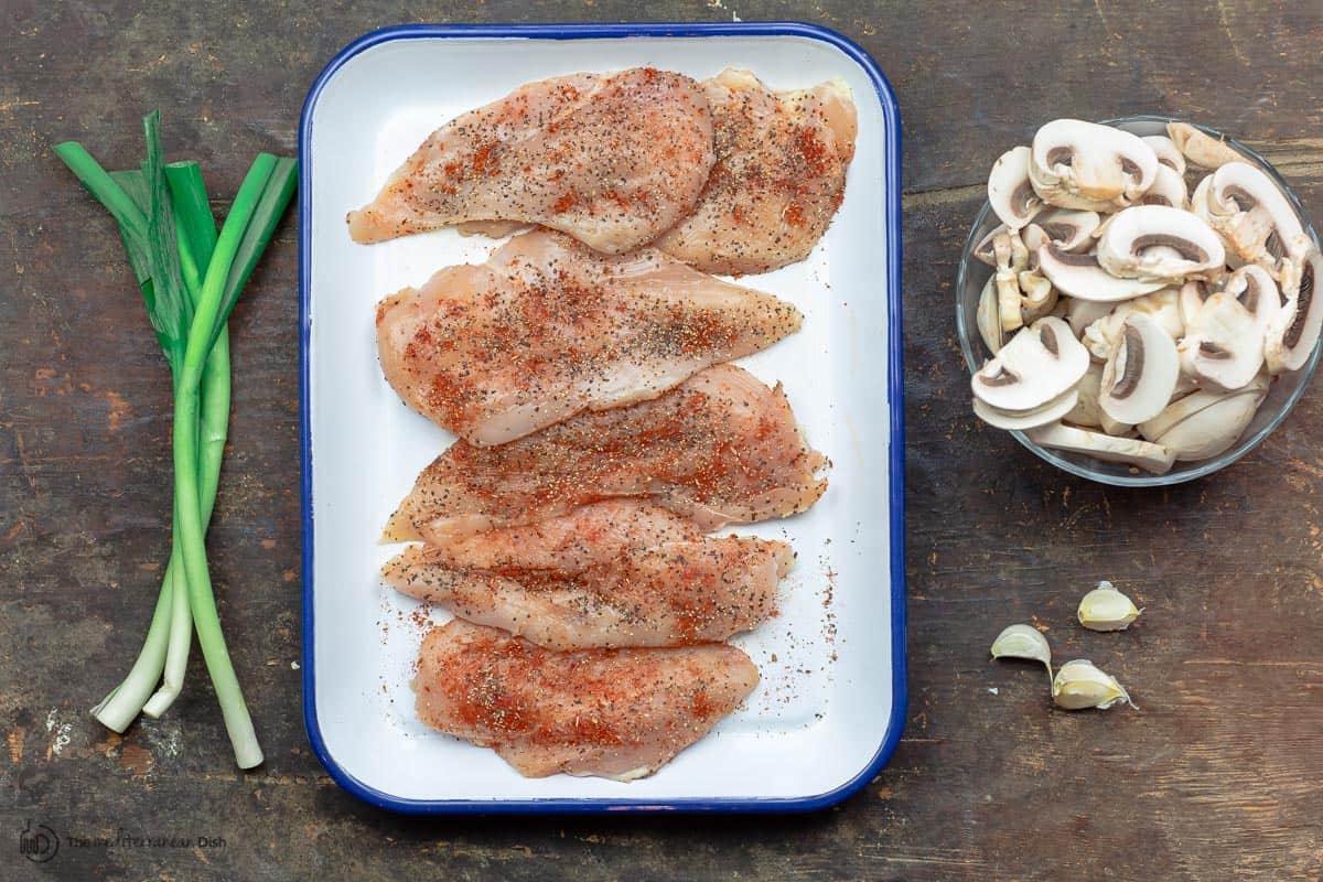 Seasoned chicken breast cutlets. A bowel of mushrooms to the side. Green onion and garlic on the table