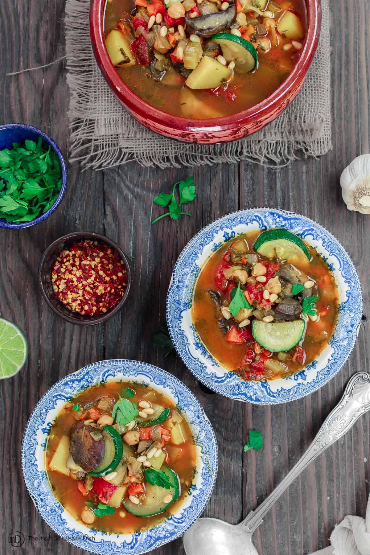 Homemade vegetable soup served in bowls with added side of fresh parsley, lime and crushed peppers