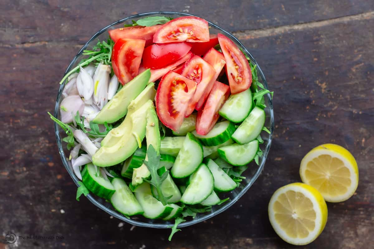 arugula salad in a large bowl with cucumbers, avocado, shallots and tomatoes. Lemons to the side 