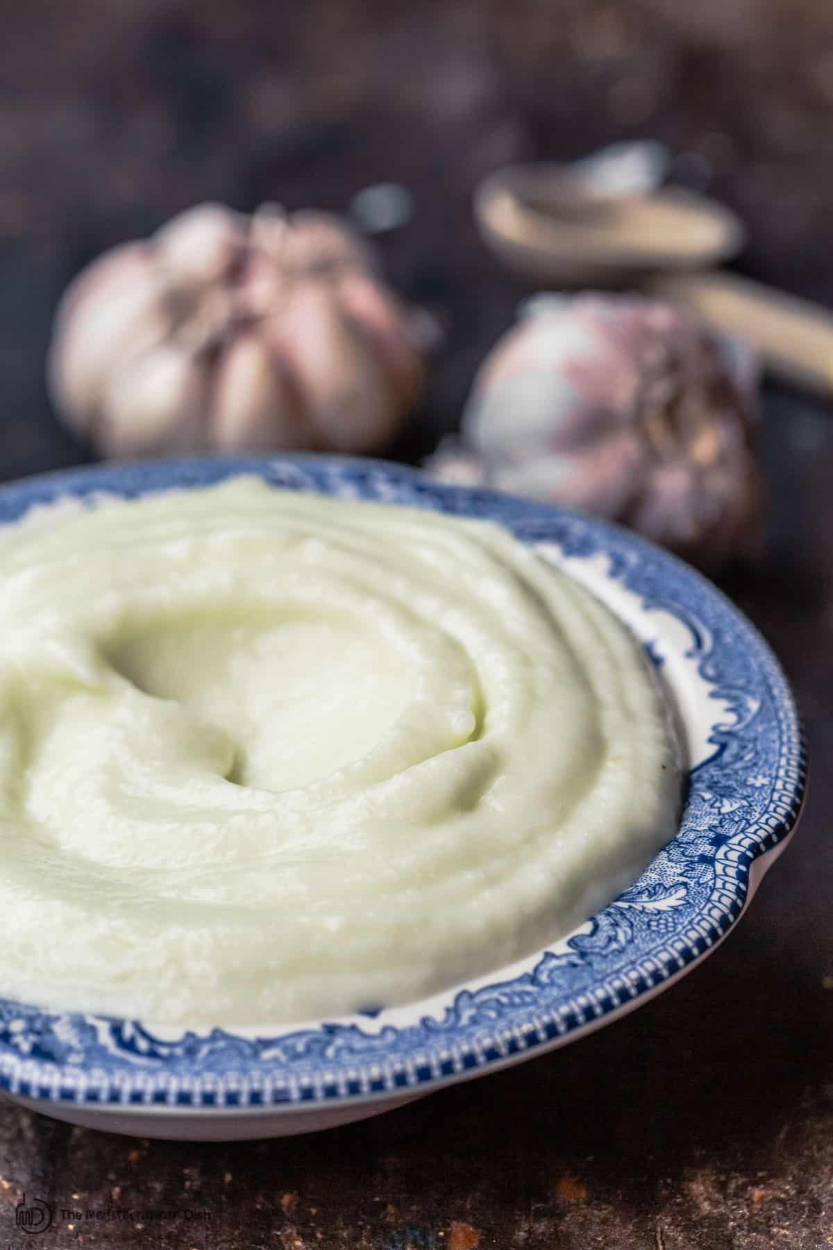 toum garlic sauce in a bowl. two heads of garlic and a spoon to the side