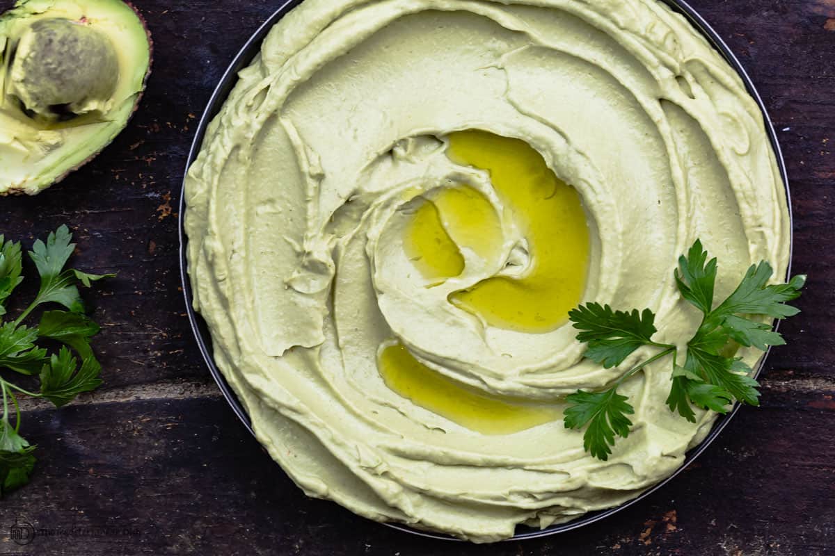 Avocado hummus dip garnished with parsley. One half an avocado to the side