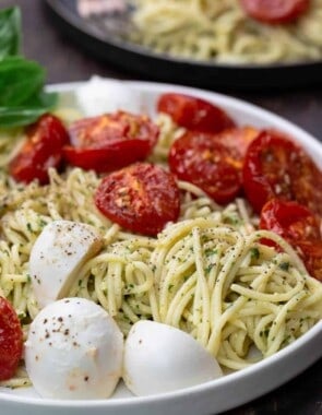 Pasta in a large bowl, tossed with pesto, tomatoes and mozzarella. A small plate to the side