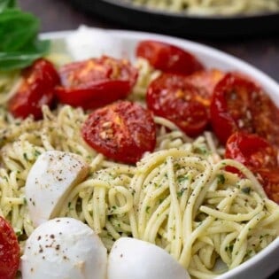 Pasta in a large bowl, tossed with pesto, tomatoes and mozzarella. A small plate to the side