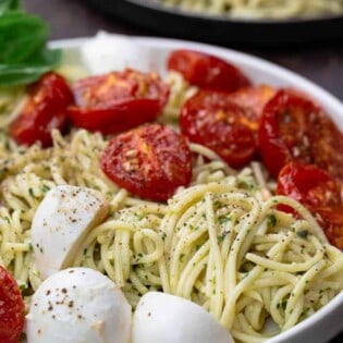 Pasta in a large bowl, tossed with pesto, tomatoes and mozzarella. A small plate to the side